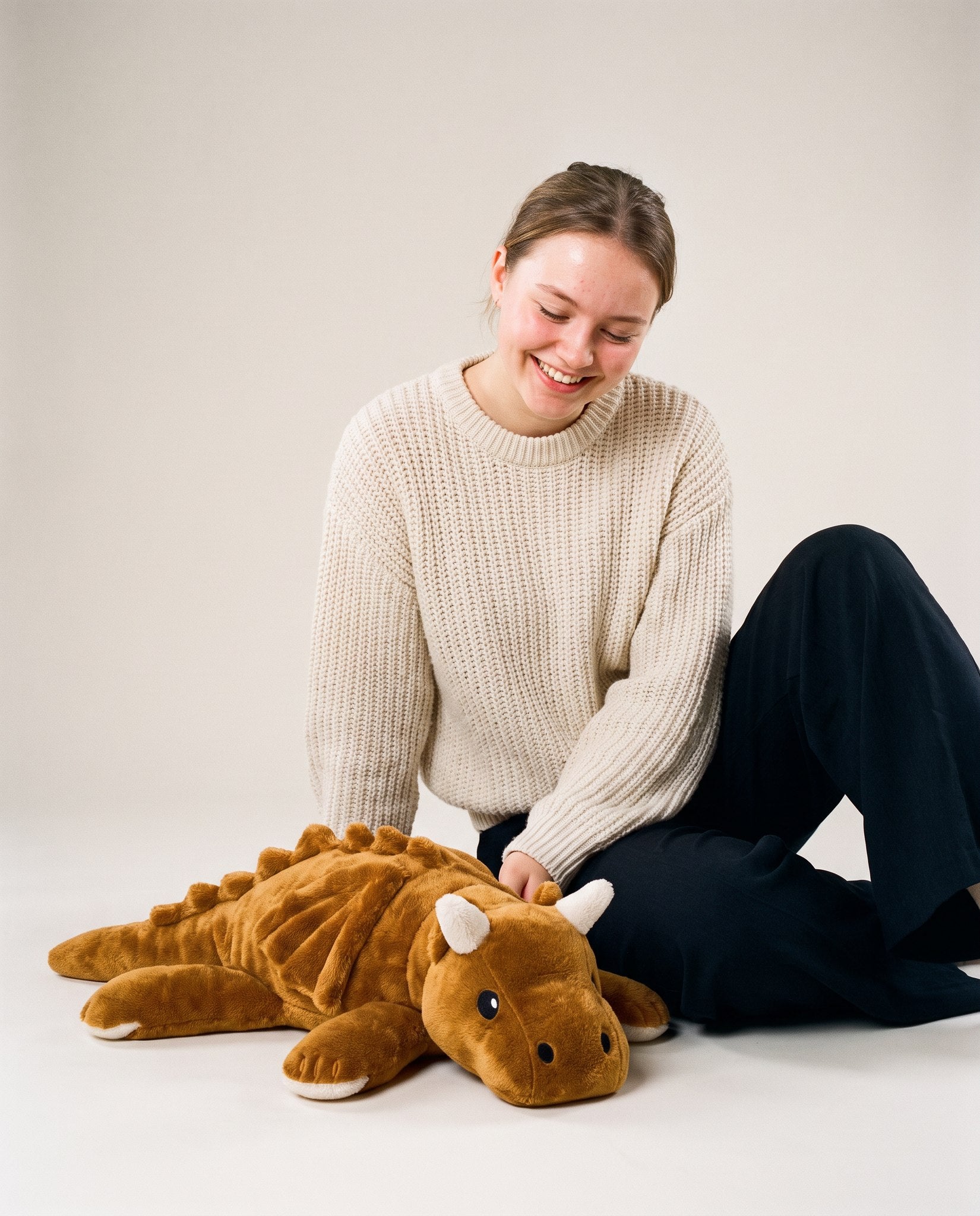 Young woman sitting on the floor smiling at the Brease Weighted Dragon 5 lb, showing its 60 cm size