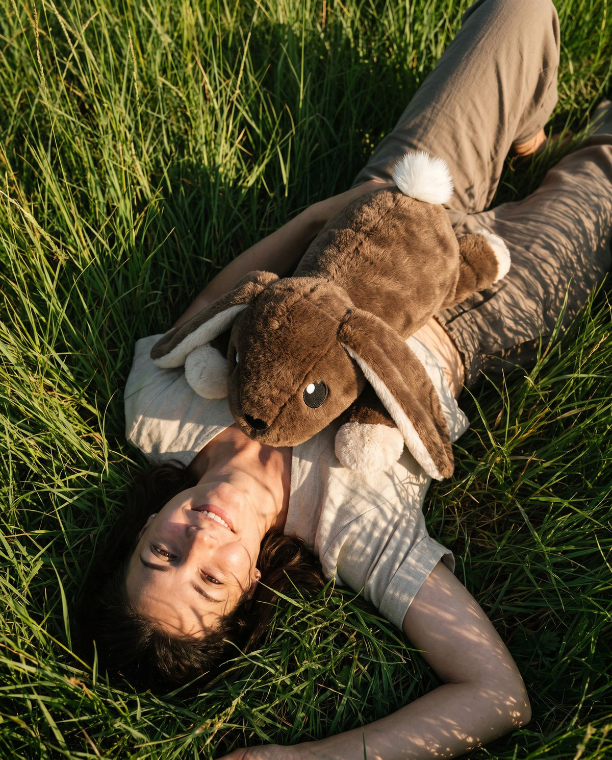 Woman lying in tall summer grass with the Brease Weighted Rabbit 5 lb resting on her chest in golden evening light